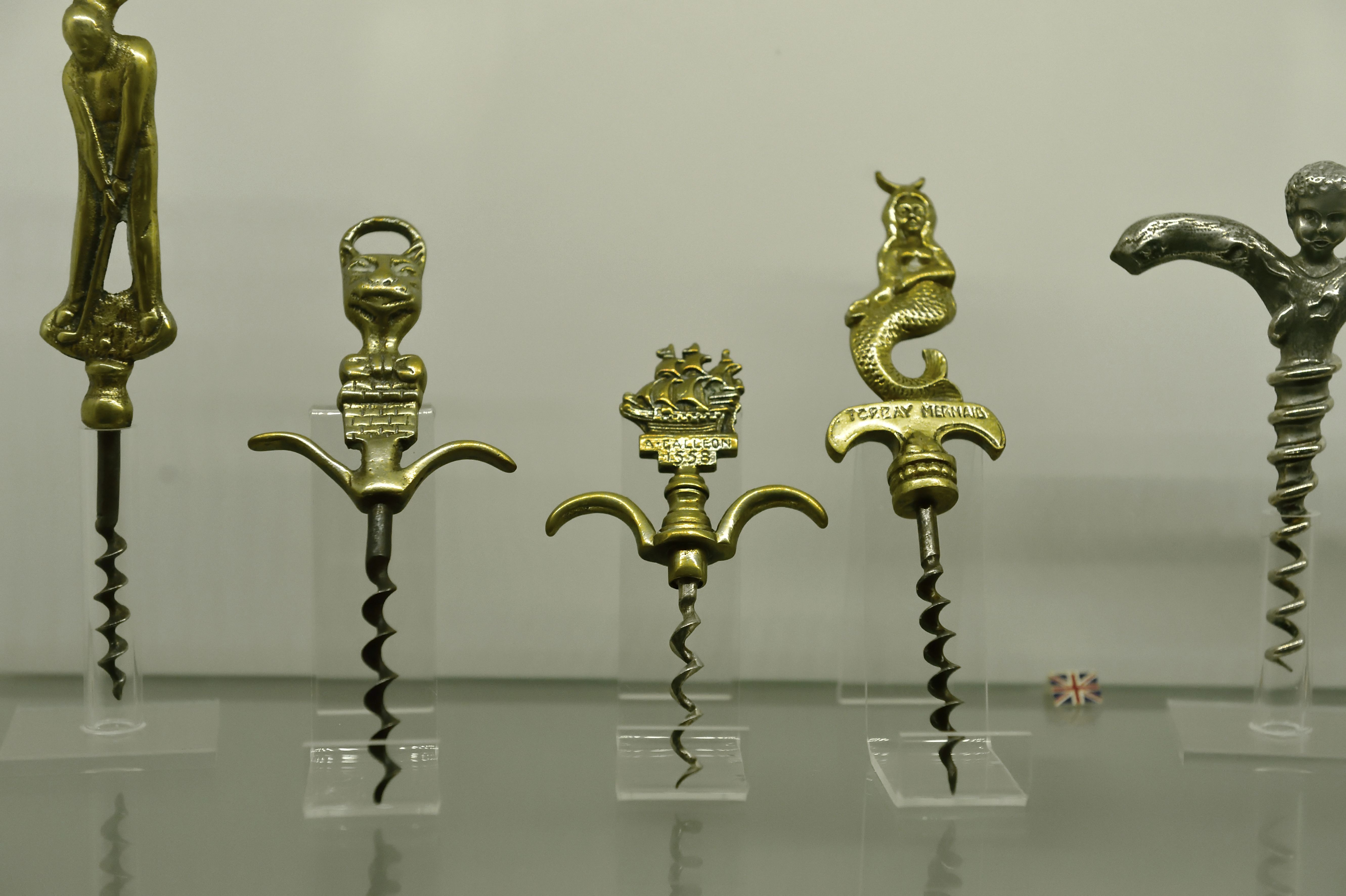 Ряд із п’яти ошатних вінтажних штопорів, виставлений на прозорій акриловій підставці на простому білому тлі. Рукоятки виготовлені з латуні та сріблястого металу різної форми, у тому числі: мах гравця в гольф. Стилізована голова кота або горгульї. корабель-галеон з позначкою "А. Галеон 1588 р."ярлик русалки "Торкі Русалка."Фігура херувима утворює ручку сріблястої спіралі.