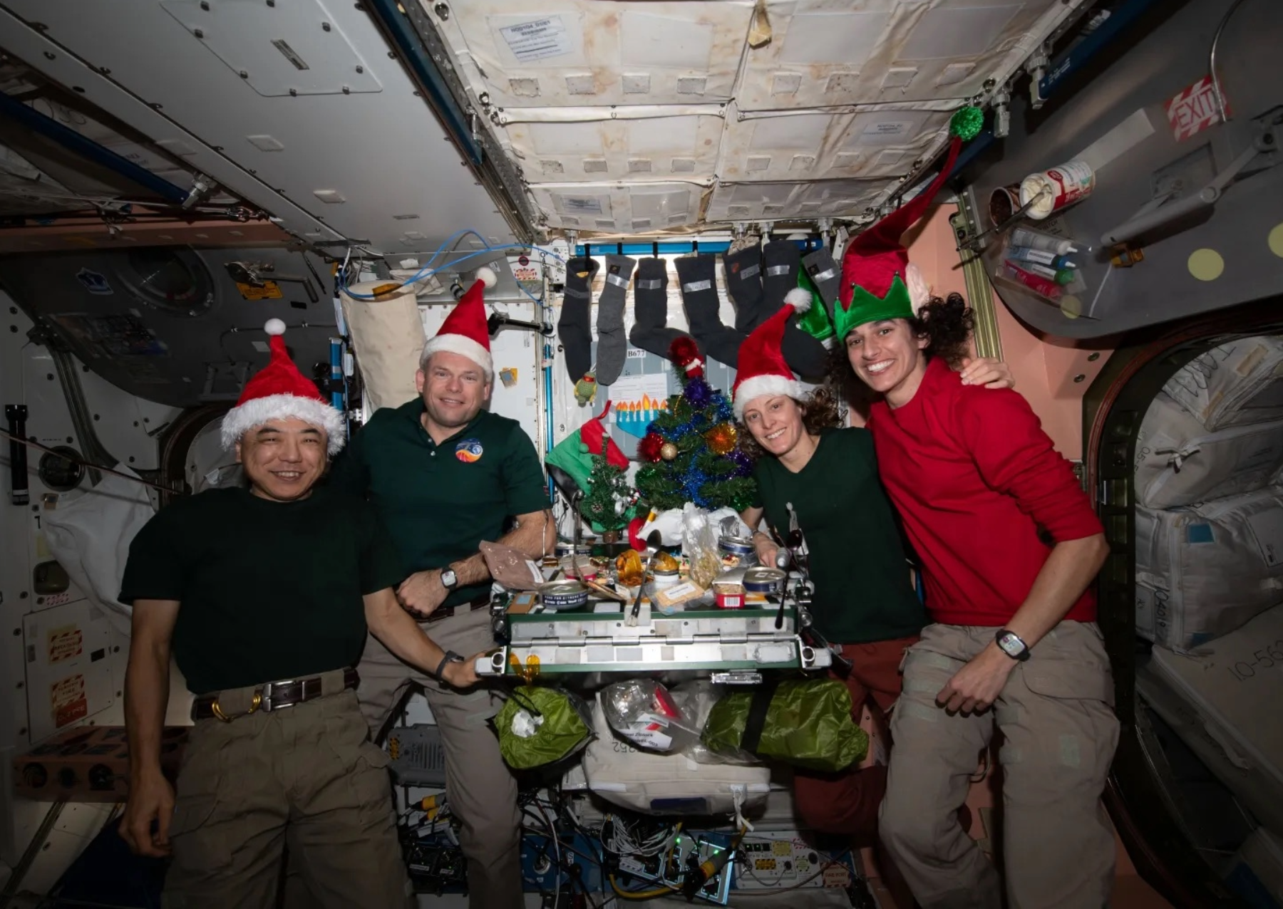 four astronauts gather around a table wearing santa hats