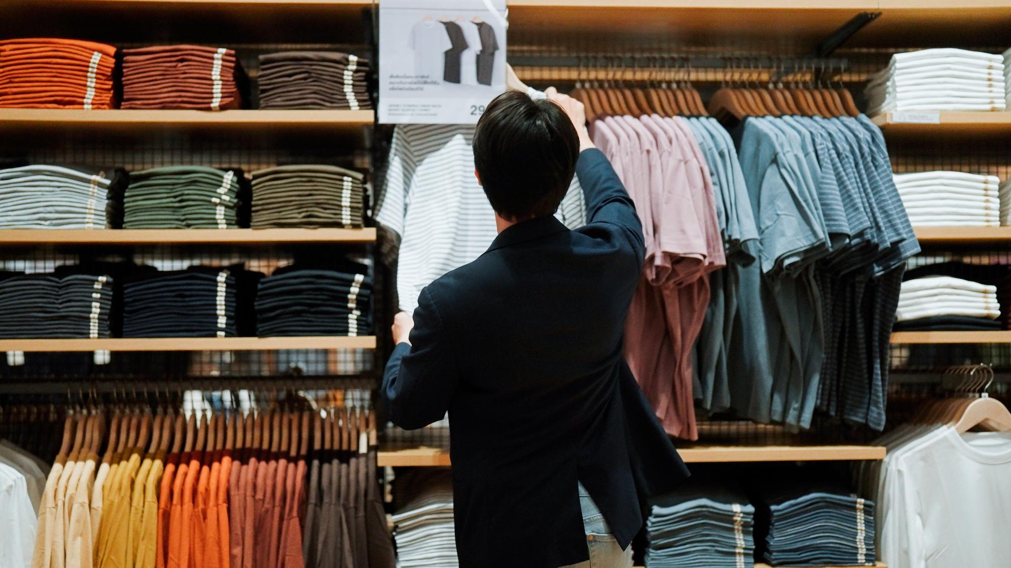 A young Asian man heading to a clothes shelf in a store before taking a closer look at a t-shirt.
