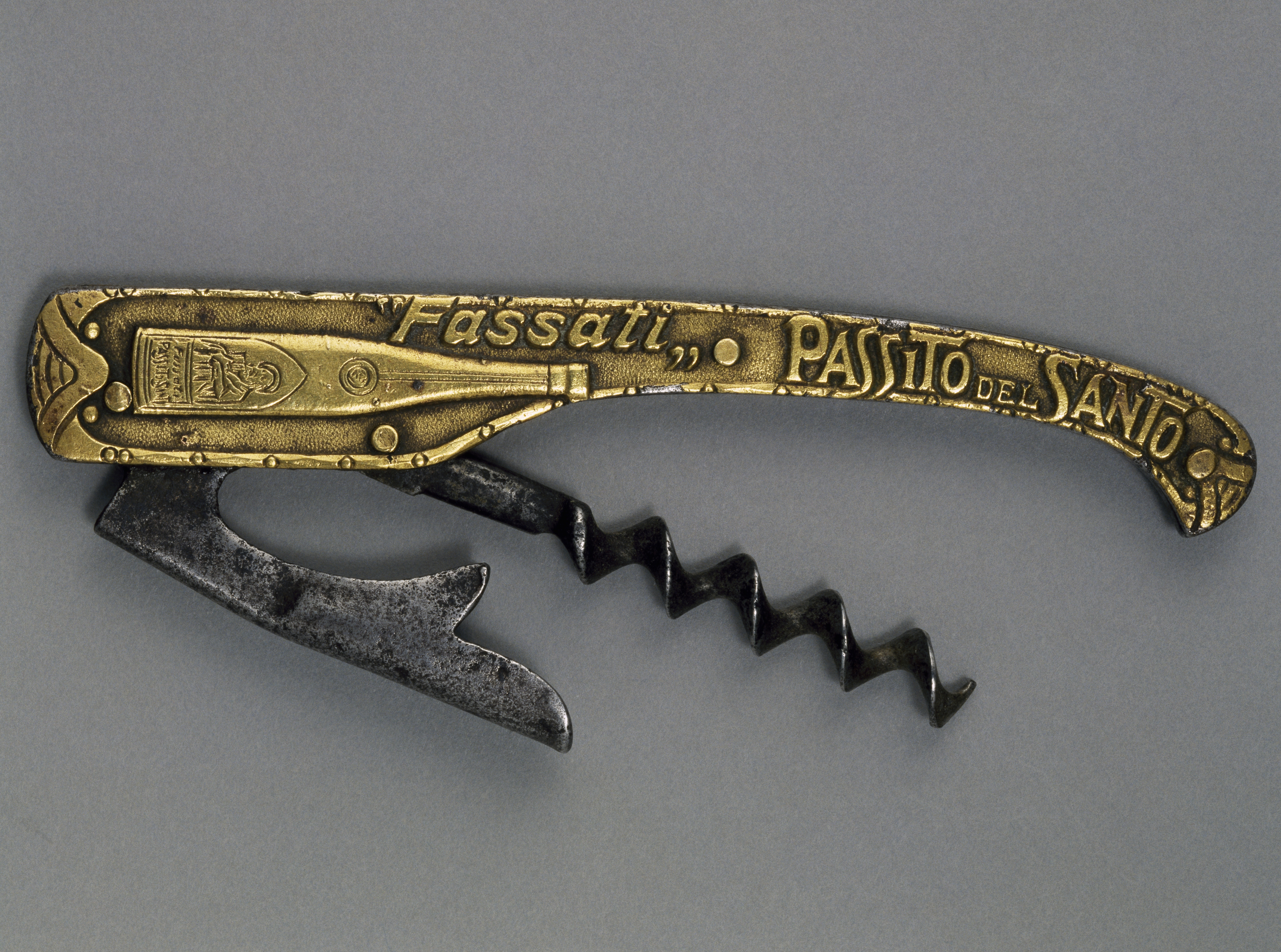 Старий складаний офіціантський штопор (дизайн Карла Вінке), зображений на нейтрально-сірому фоні. Ручка виготовлена ​​з металу золотистого тиснення з декоративним текстом "Фасаті Пасіто дель Санто" І ілюстрація пляшки вина. Від ручки вигнутий темний металевий спіральний черв’як і зубчастий важіль сріблястого кольору, що відображає ранню механічну конструкцію портативних відкривачок для вина.