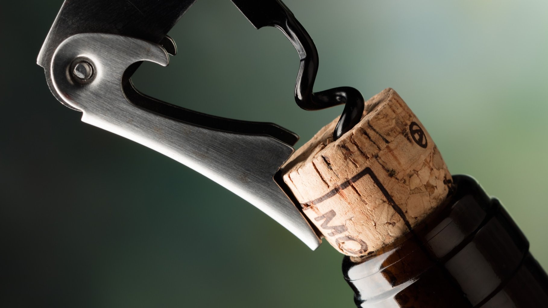 A close-up, angled shot of a silver waiter's corkscrew in the process of opening a wine bottle. The metal lever of the corkscrew is braced against the lip of the dark glass bottle, while the black spiral "worm" is drilled deep into the center of the natural wood cork. The background is a soft, out-of-focus gradient of green and grey.