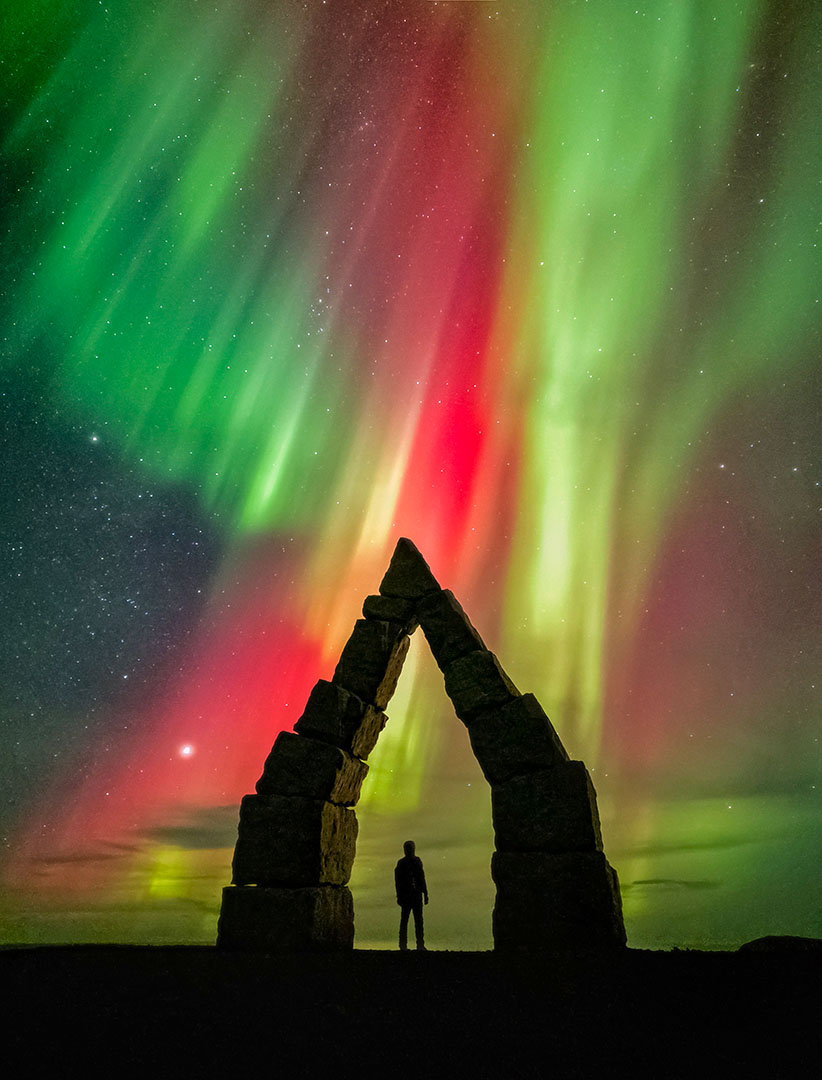 a man stands under an arch while a red and green aurora dances in the night sky