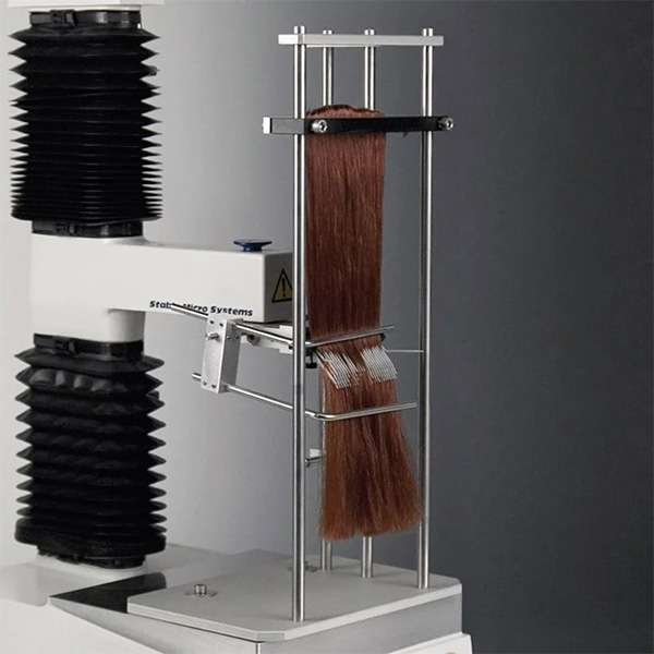 A laboratory testing rig featuring a long tress of auburn hair held vertically within a metal frame. A motorized arm equipped with a comb is positioned to move through the hair sample, used for measuring combing force and hair manageability.