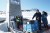 The concrete entrance of the Svalbard Global Seed Vault sits nestled in a snowy mountainside under a clear blue sky. In the foreground, several people are gathered on a metal walkway near the vault's open door. A stack of blue and black storage crates, labeled with the