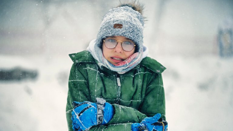 A young person bundled in a green winter jacket, gray knit hat, and blue gloves is standing outside in the snow. They are shivering, with their arms crossed tightly and a nervous or cold expression, with their tongue slightly visible.