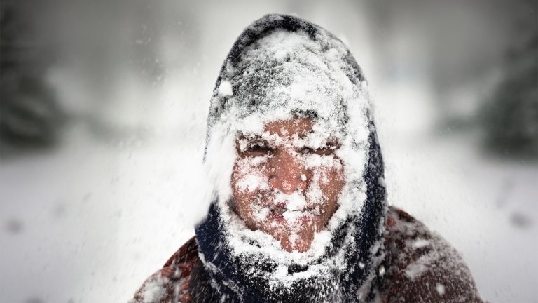 A person bundled up in a hooded jacket and covered in snow is looking toward the camera in a snowy, blurry outdoor environment. The person's face is mostly obscured by a thick layer of snow and frost, with only the area around the eyes and nose showing exposed skin.