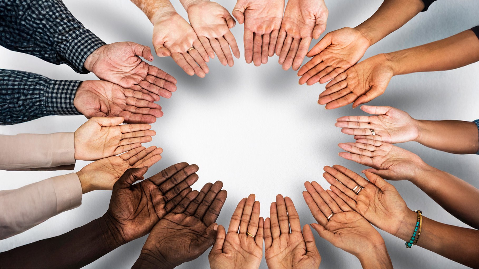 An overhead view shows a group of diverse hands, with varying skin tones, arranged in a circle against a plain white background. The palms are open and facing upward, with some hands overlapping, creating a sense of unity and community. Some individuals wear rings or a beaded bracelet, and the sleeves of various shirts—plaid, denim, and solid colors—are visible at the edges of the frame.
