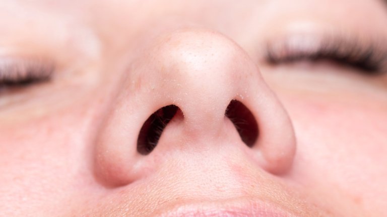 A macro, close-up photograph of the bottom of a human nose, clearly showing the two nostrils and the texture of the surrounding skin. The eyes are closed in the background.