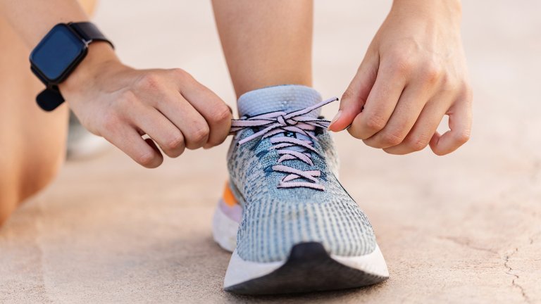 A close-up photograph of a person’s hands tying the light pink laces of a blue and gray patterned running shoe. The person is wearing a black smartwatch on their left wrist, and the background consists of a blurred, light-colored concrete surface.