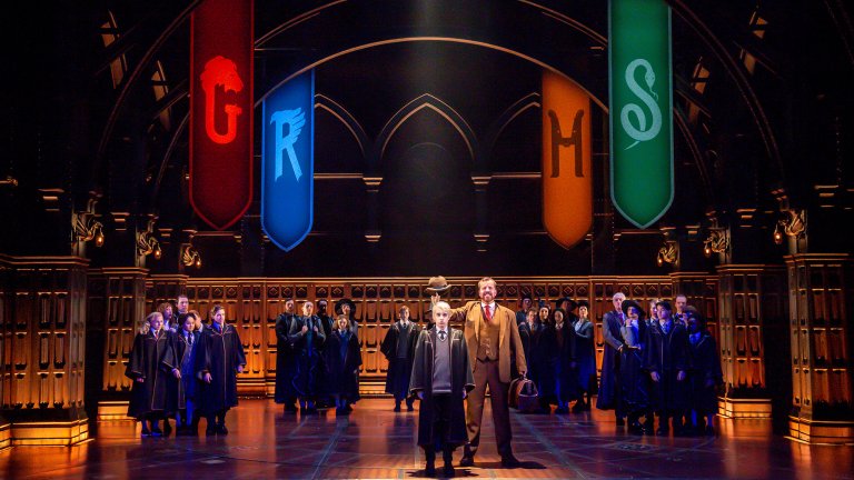A vibrant stage production of Harry Potter and the Cursed Child. In the center, a young wizard is being sorted under a floating hat by a man in a brown suit. Four large, illuminated banners representing the Hogwarts houses—Gryffindor (red), Ravenclaw (blue), Hufflepuff (yellow), and Slytherin (green)—hang from the gothic arches of the Great Hall, while students in robes watch from the sides.