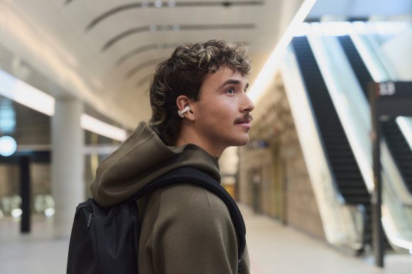 A millenial with a mustache wandering around in public transport showing off a Sony Linkbud Clip open earbud in greige