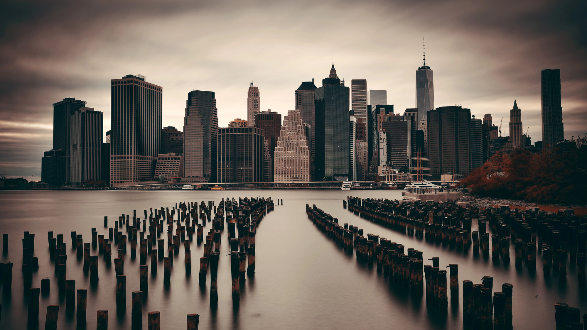 An atmospheric, long-exposure photograph of the New York City skyline as seen from across a river. In the foreground, dozens of weathered wooden pilings from an old pier protrude from the smooth, glass-like water, leading the eye toward the dense cluster of skyscrapers in Lower Manhattan. The sky is filled with heavy, dark, brooding clouds, and the image has a desaturated, somber color palette with hints of autumn foliage on the right bank.
