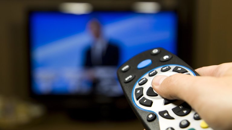 Hand holding TV remote in front of out of focus TV screen