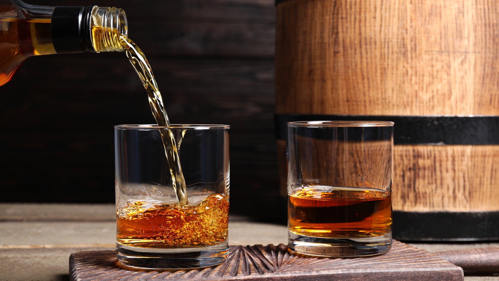 Pouring whiskey into glass from bottle near wooden barrel on table