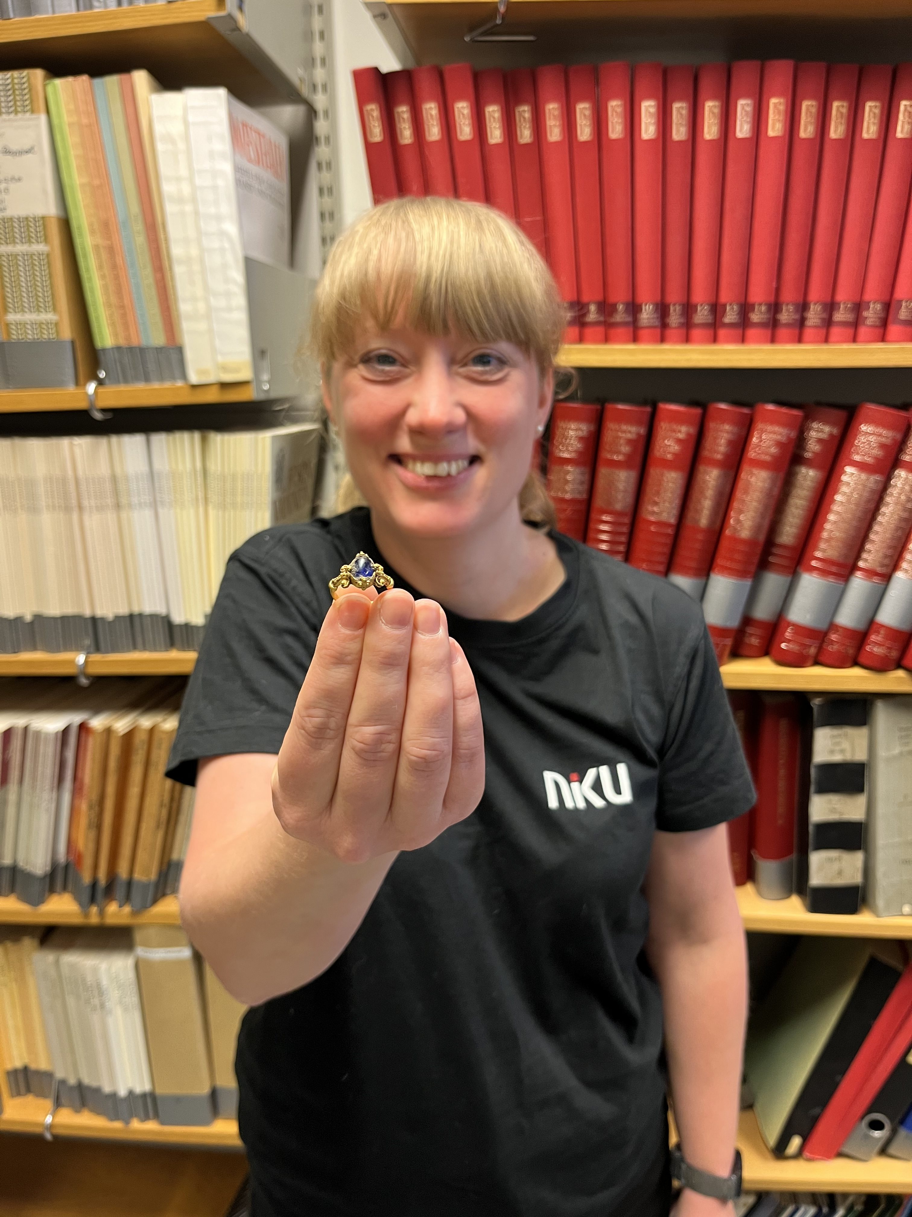 a woman holds up a medieval ring while standing in front of a bookshelf