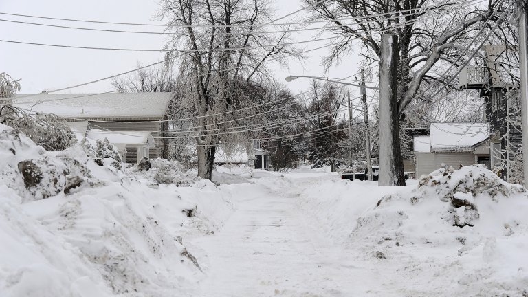Snowed in suburban street. Piles of snow are on either side of a snowy road. A house and telephone wires are seen in the background.