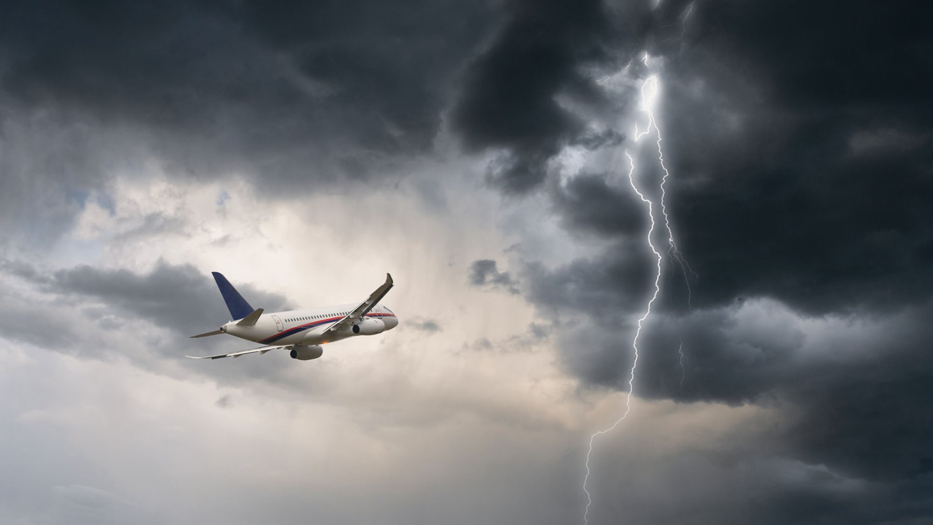 Commercial airplane flying in stormy weather. A lightning strike illuminates the right side of the image as a plane flies to the left.