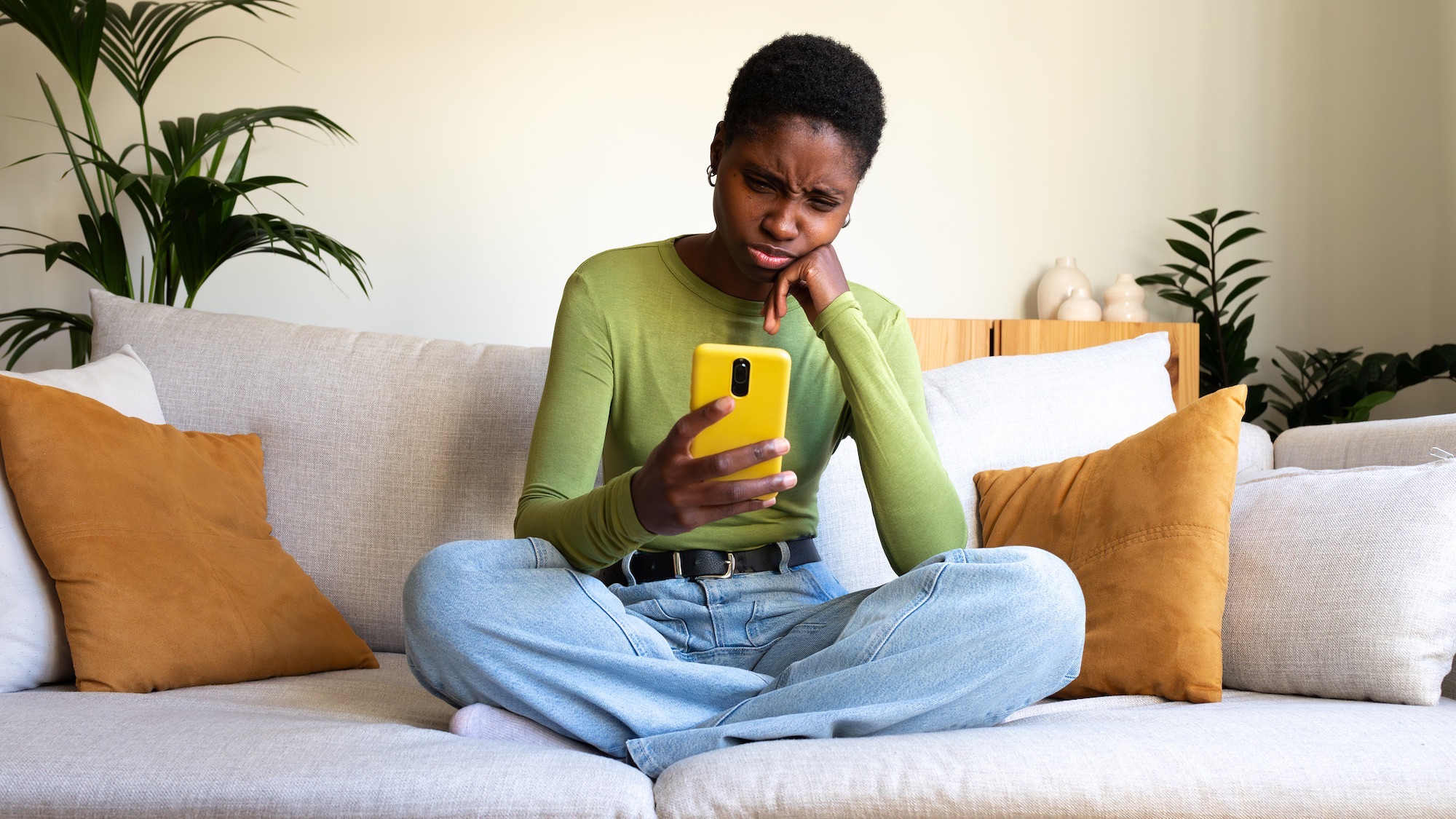 Young woman sitting on a sofa and looking at a smartphone with a confused expression