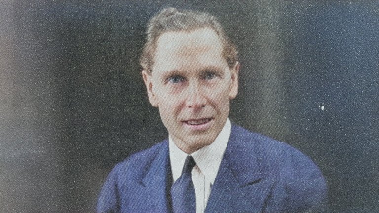 A vintage-style colorized portrait of a middle-aged man with light hair combed back, wearing a blue suit jacket, white shirt, and dark tie. He faces the camera with a neutral expression. The background is a uniform, grainy gray, and the image retains visible texture and noise consistent with an old printed photograph.