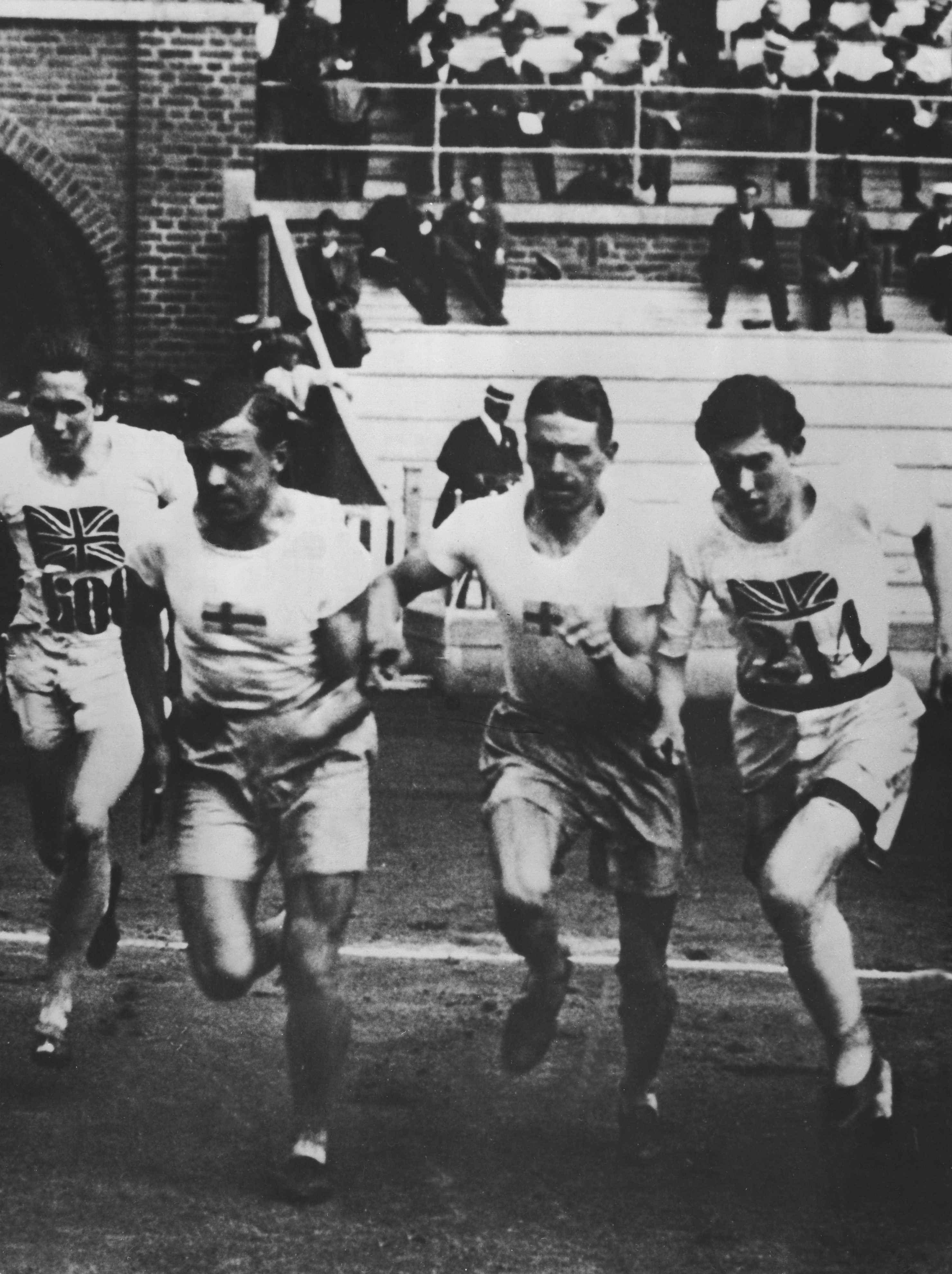 A black-and-white action photograph from the 1912 Stockholm Olympics showing four men competing in a middle-distance track event. Philip Noel-Baker is on the far right, wearing a white jersey with a Union Jack and the number 214, captured mid-stride alongside competitors from Finland and other nations. A crowd of spectators in hats is visible in the background bleachers.