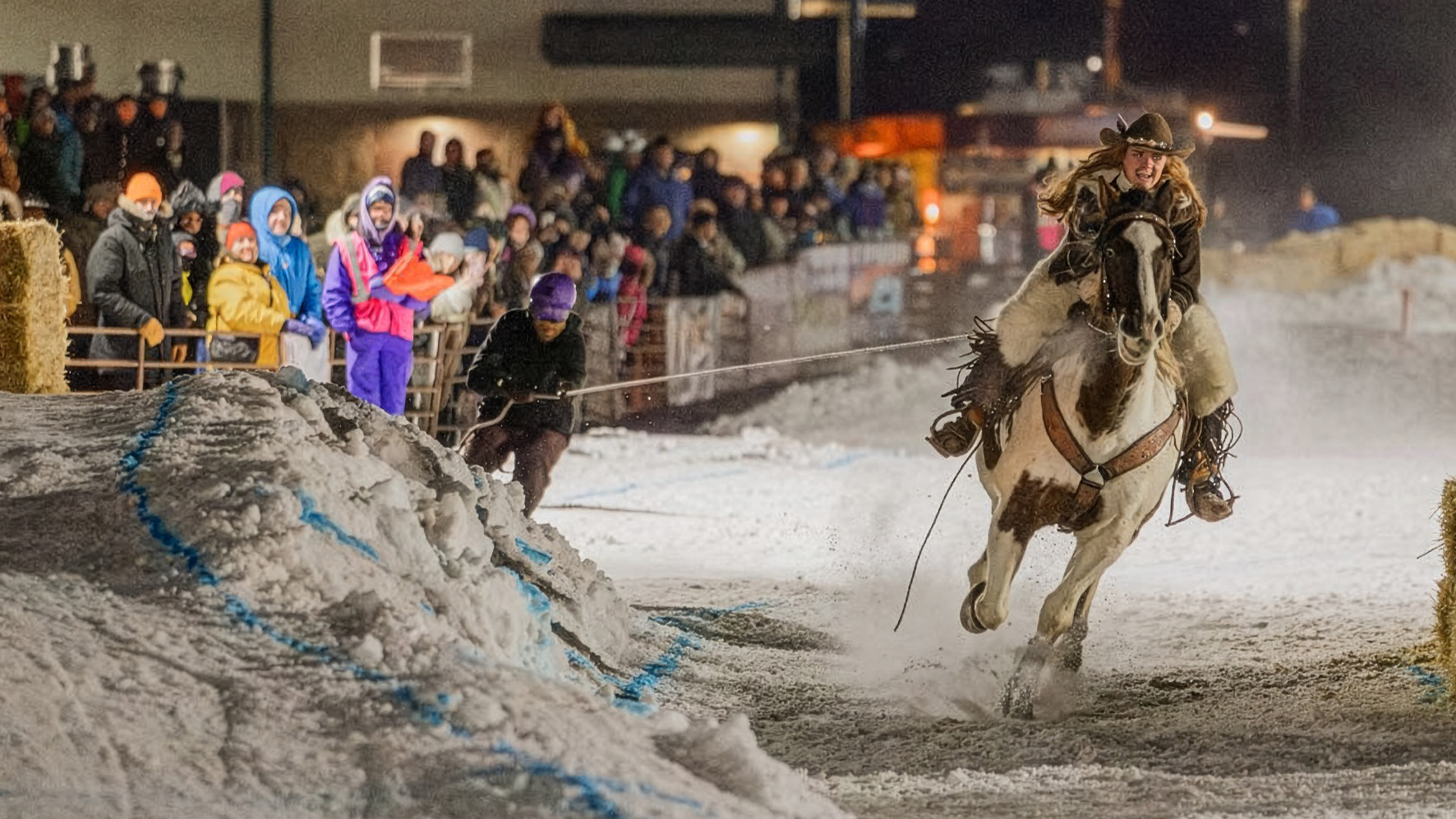 An action-packed, night-time shot of a skijoring competition. A person on skis is being pulled at high speed by a galloping, brown-and-white paint horse. The rider, a woman in a cowboy hat and western gear, leans forward as the horse kicks up snow. In the background, a large crowd of spectators in winter clothing watches from behind a fence, illuminated by bright stadium lights. The foreground features snowy mounds and hay bales marking the course.