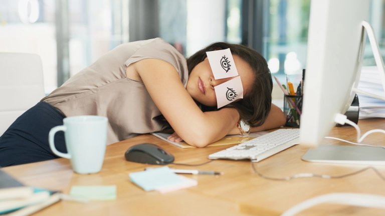 A young woman slumped over her desk in an office, fast asleep on her arms. To humorously hide that she is sleeping, two pink sticky notes with cartoon eyes drawn on them are stuck over her closed eyelids. Her workspace includes a computer monitor, keyboard, mouse, and a light blue coffee mug.