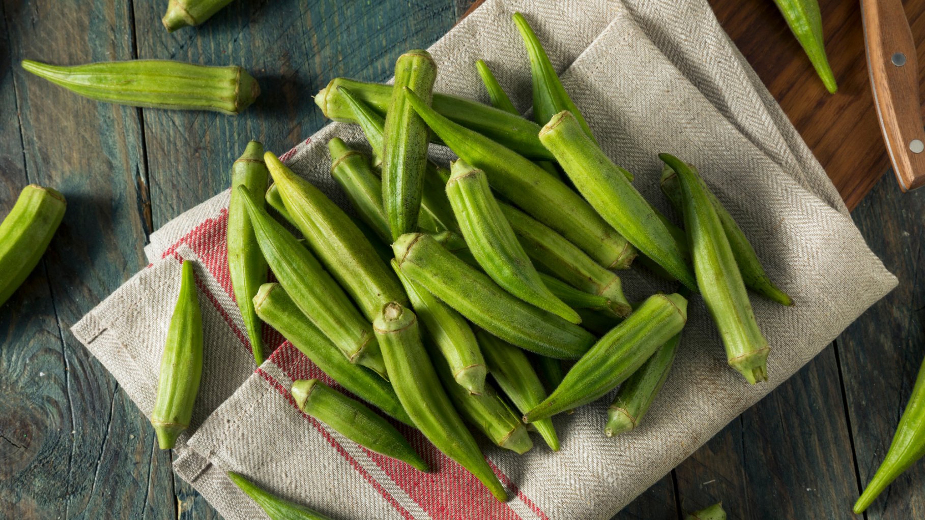 A top-down photograph of a pile of fresh, green okra pods resting on a rustic, beige linen napkin with a red striped border. The pods are scattered across the fabric and a weathered wooden tabletop, with the wooden handle of a kitchen knife visible in the upper right corner.