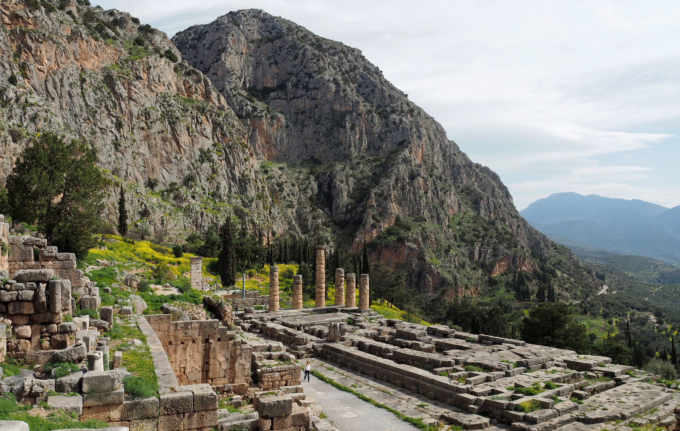 A wide-angle landscape photograph of the ancient ruins of the Temple of Apollo in Delphi, Greece. The stone foundations and several standing Doric columns are situated on a steep mountain slope under a bright, partly cloudy sky, overlooking a lush green valley.