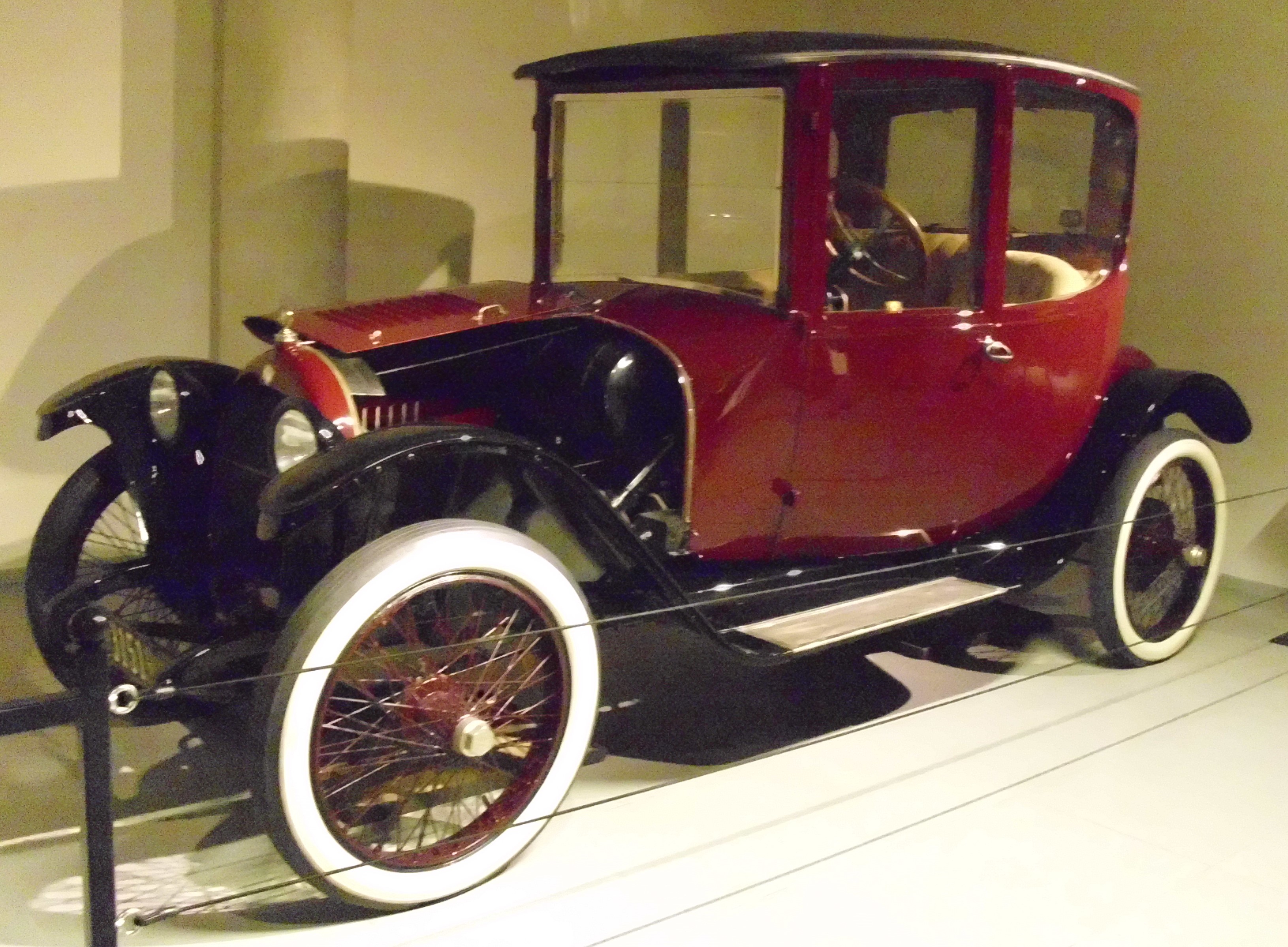 A red and black 1917 Woods Dual Power antique car with white-walled tires, displayed in a museum. The car features a tall, glass-enclosed cabin and a split hood design.