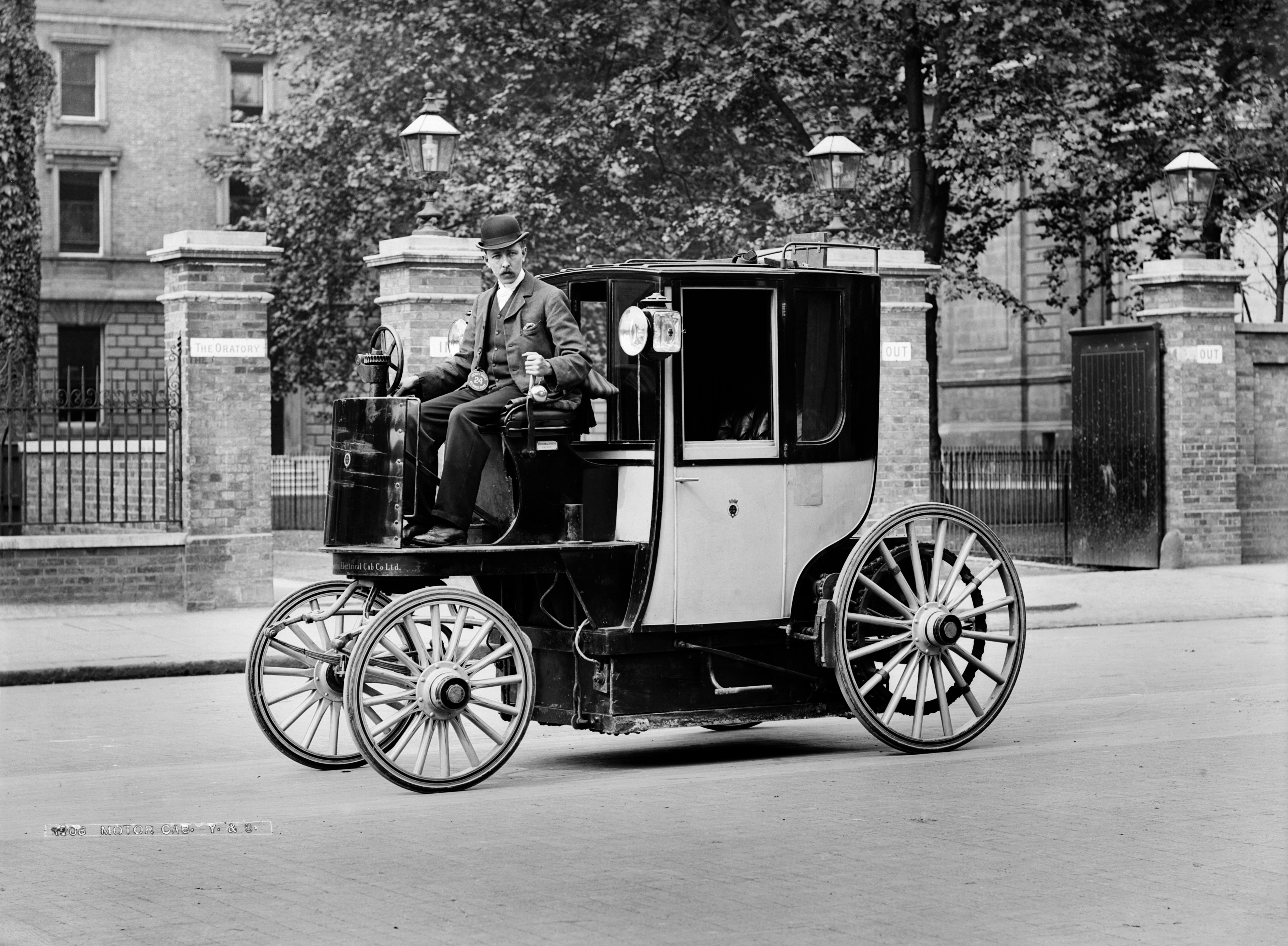 A black and white historical photograph of a Bersey Electric Cab, a vintage electric taxi, parked on a London street in front of the Brompton Oratory.

The vehicle features a unique design with a large, enclosed passenger carriage at the rear and an open, elevated driver’s seat at the front. A man wearing a three-piece suit and a bowler hat sits at the steering wheel, which is a vertical column. The cab has four large, light-colored wooden-spoke wheels and two lanterns mounted near the passenger door. In the background, brick pillars with "IN" and "OUT" signs flank a gated entrance, and a large stone building is visible behind a row of trees.