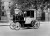 A black and white historical photograph of a Bersey Electric Cab, a vintage electric taxi, parked on a London street in front of the Brompton Oratory.
The vehicle features a unique design with a large, enclosed passenger carriage at the rear and an open, elevated driver’s seat at the front. A man wearing a three-piece suit and a bowler hat sits at the steering wheel, which is a vertical column. The cab has four large, light-colored wooden-spoke wheels and two lanterns mounted near the passenger door. In the background, brick pillars with