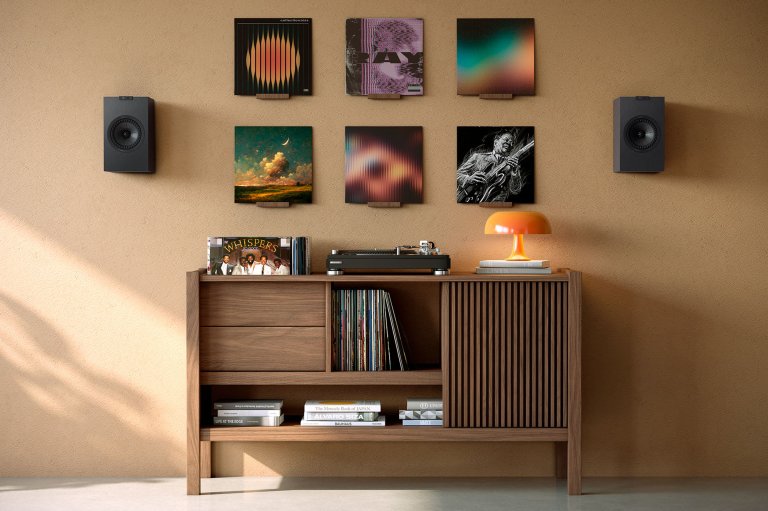 Two satin black KEF Q4 Meta on-wall speakers flanking vinyl album artwork hung over a credenza with a turntable on it.