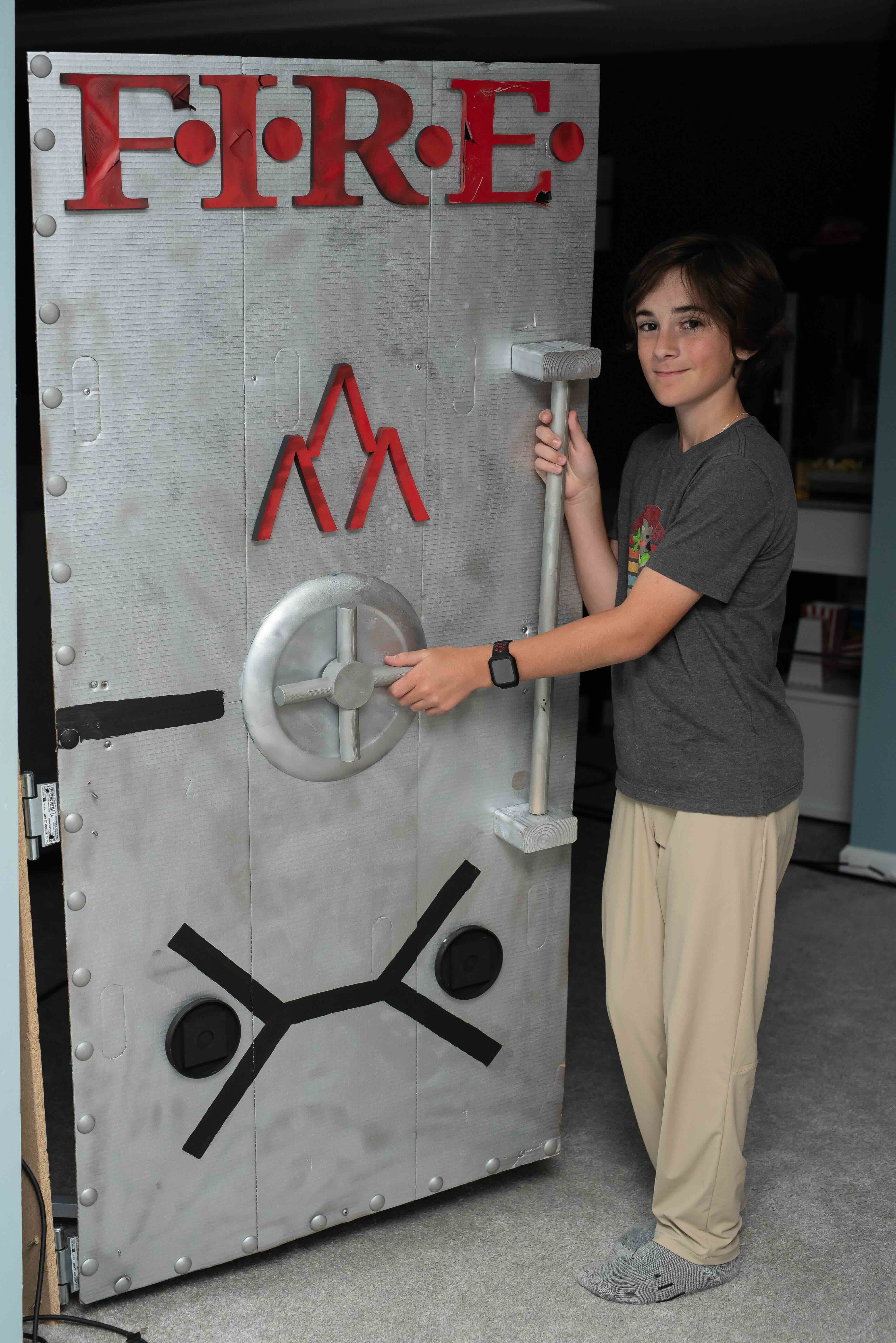 A medium shot of a smiling teenage boy with brown hair, standing next to a large, custom-built prop door designed to look like a metallic vault or industrial hatch.
The door is painted silver with a weathered texture and features several distinct elements:
The word "FIRE" in large, bold red letters at the top.
A red mountain-like logo in the center.
A silver circular wheel handle and a vertical grab bar.
A black symbol at the bottom resembling a stylized person or a schematic.
Silver decorative rivets lining the left and bottom edges.
The boy is wearing a grey t-shirt, tan pants, and grey socks, with one hand on the wheel and the other on the grab bar. The background appears to be an indoor room with light blue walls.