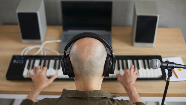 Professional male musician creates new song for music album by playing synthesizer in studio. Top view at angle on head of bald man in headphones who sits at microphone and presses keys of synthesizer