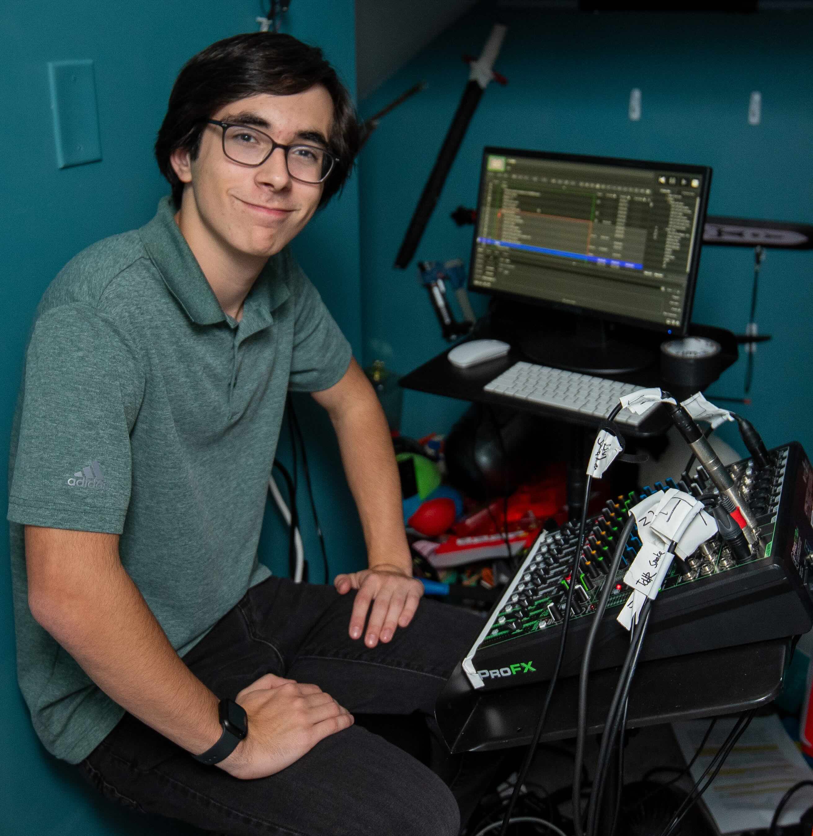 A young man with glasses and a green polo shirt sits in front of a technical workstation, featuring a computer monitor displaying audio editing software and a Mackie ProFX mixing console with labeled cables.