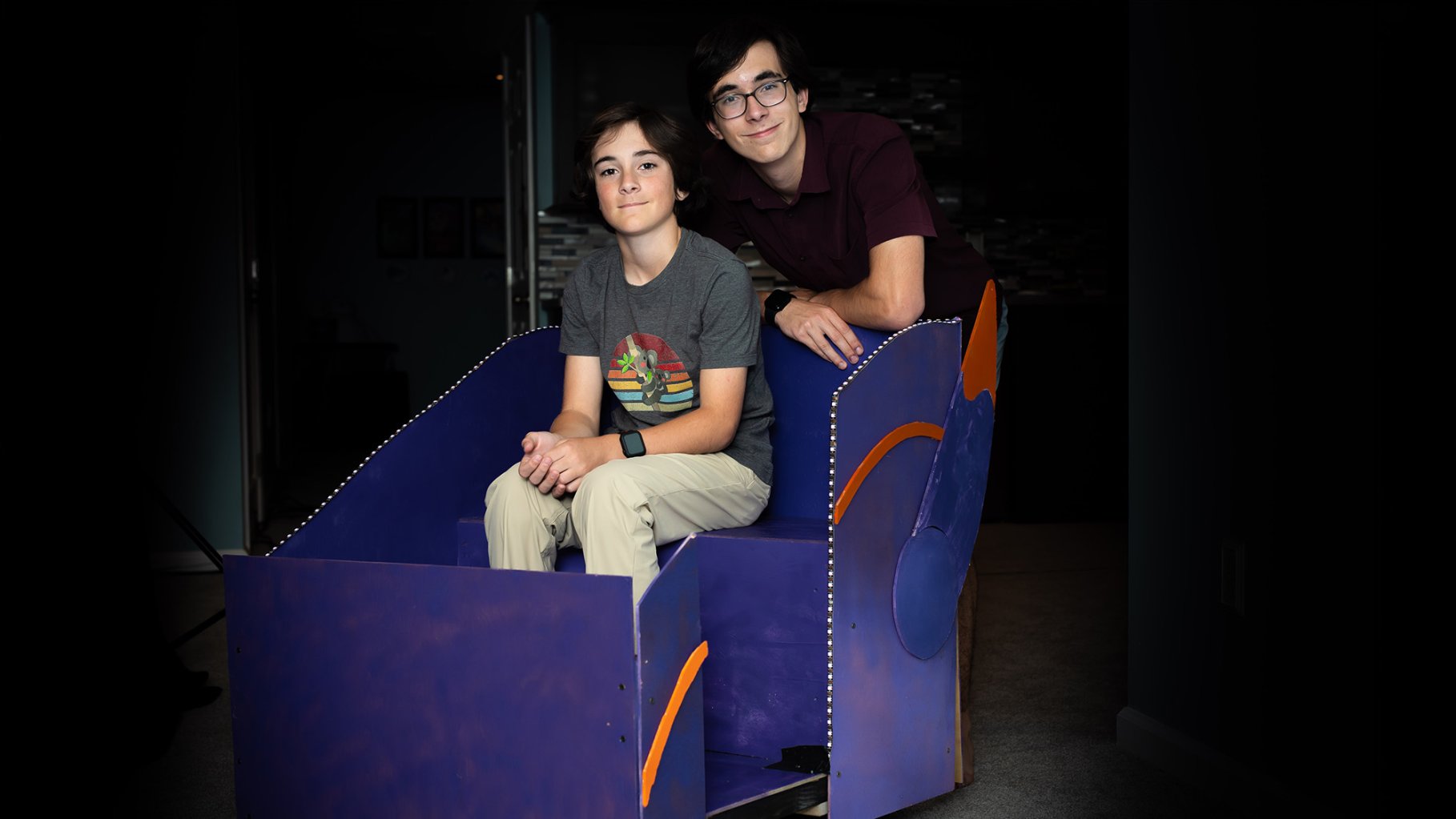 A portrait of two brothers, Nico and Matteo Mucchetti, posing with their homemade, full-sized replica of a purple ride vehicle from Disney's "Big Hero 6" attraction. The younger brother sits inside the purple wooden structure, which is detailed with orange accents and a string of LED lights along the edge, while the older brother leans over the back. They are positioned in a dimly lit room, creating a dramatic spotlight effect on them and their project.