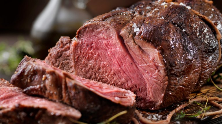 A close-up photograph of a thick-cut roast of venison, sliced to reveal a deep pink, juicy interior. The meat is tied with butcher's twine and has a dark, seasoned crust. It is garnished with coarse sea salt and fresh herbs, resting on a rustic wooden carving board with a blurred kitchen background.