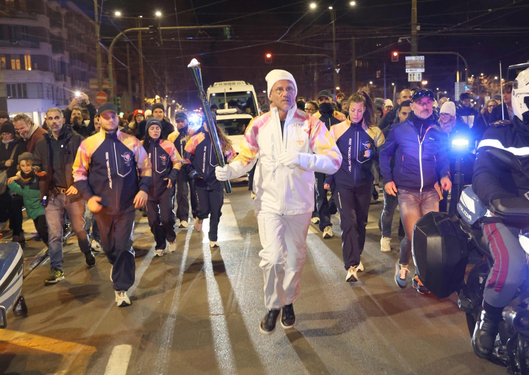 a man in a white snow suit and winter hat carries a torch through the street