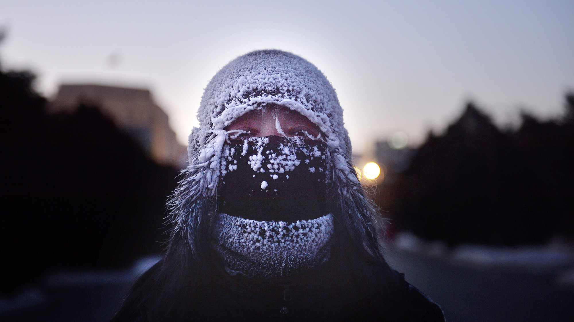 A person bundled in heavy winter gear, including a frost-covered knit hat and a black face mask, with ice clinging to their eyelashes and the fabric. The background shows a dim, icy landscape at dusk with a soft glow from a distant light.