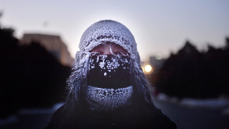 A person bundled in heavy winter gear, including a frost-covered knit hat and a black face mask, with ice clinging to their eyelashes and the fabric. The background shows a dim, icy landscape at dusk with a soft glow from a distant light.