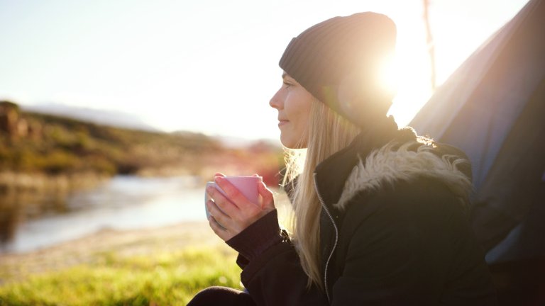 A side-profile photograph of a woman in a black winter coat and beanie, holding a pink mug with both hands. She is outdoors at sunrise or sunset, with a bright sun flare behind her head, a blue tent to her right, and a river landscape in the background.