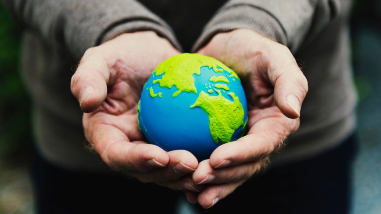 A close-up photograph of a person's cupped hands holding a small, vibrant blue and green globe, symbolizing care and responsibility for the planet. The background is softly blurred, focusing all attention on the fragile Earth resting in the palms.