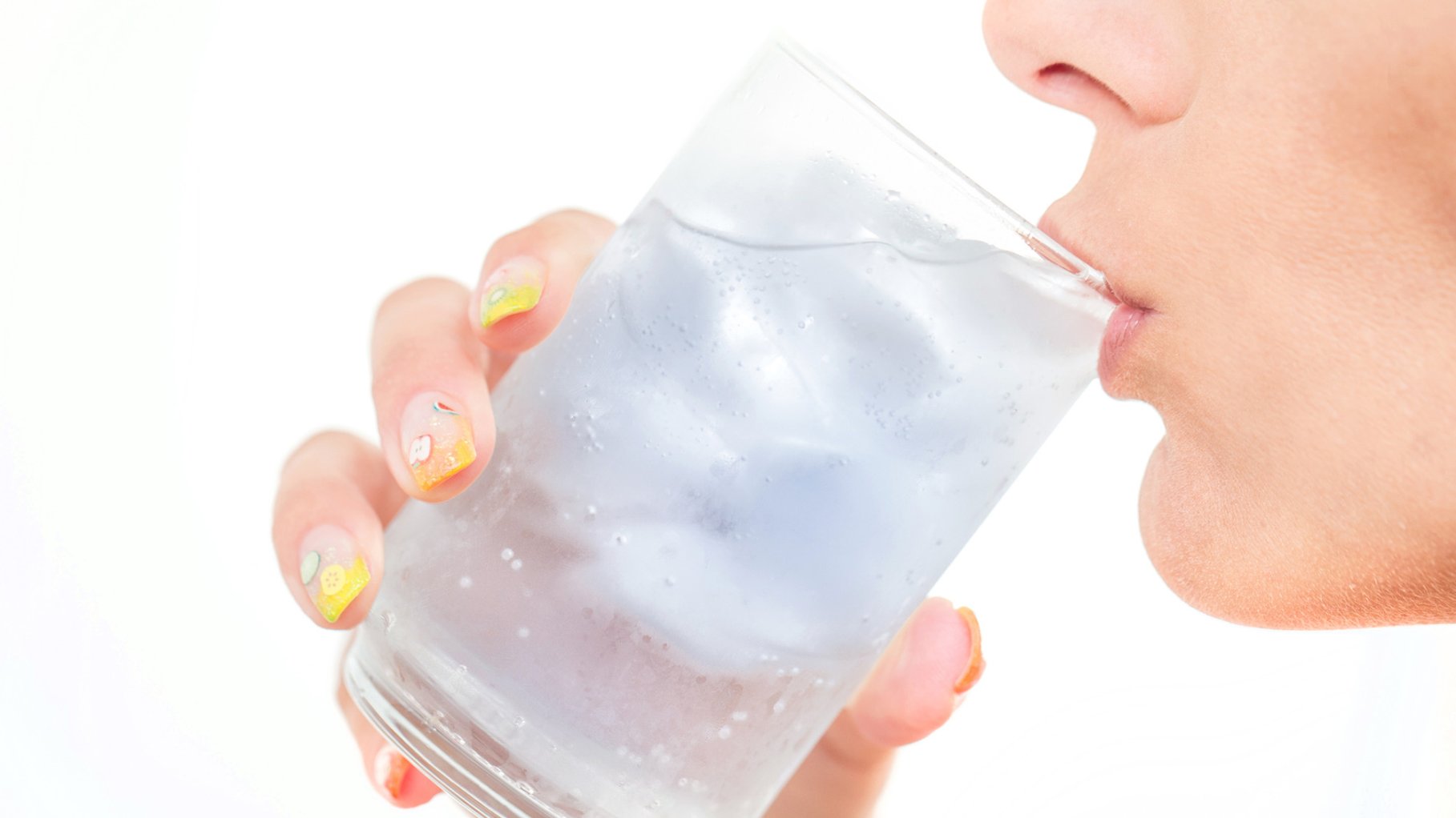 A bright, high-key close-up photograph of a woman drinking from a clear glass filled with ice and water. The focus is on the glass and her hand, which features decorative nail art with small fruit patterns.