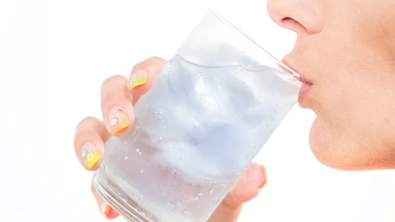 A bright, high-key close-up photograph of a woman drinking from a clear glass filled with ice and water. The focus is on the glass and her hand, which features decorative nail art with small fruit patterns.