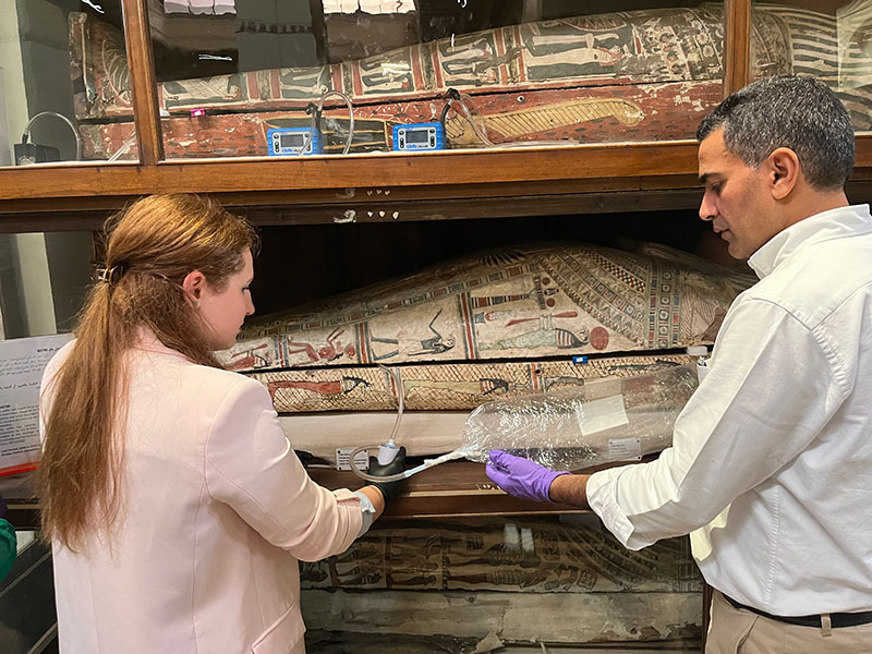 A medium shot of two researchers, a woman in a pink blazer and a man in a white button-down, working in a museum or archive. They are carefully sampling the air from inside a middle-tier Egyptian sarcophagus using a thin tube connected to a clear collection bag. The sarcophagus is intricately painted with hieroglyphs and figures, and more coffins are visible on the shelves above and below it. Small blue digital monitoring devices are positioned on the shelf above the main sarcophagus.