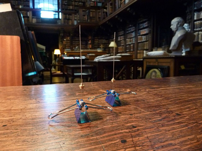 A close-up, low-angle shot of two small air-sampling needles standing upright on a polished wooden table. Each needle is held in place by a blue metal binder clip. The background is a large, dim, multi-story historical library with floor-to-ceiling bookshelves, rolling ladders, and a white marble bust on a pedestal to the right. The focus is sharp on the sampling equipment in the foreground, with the grand library blurred into a warm, scholarly atmosphere.