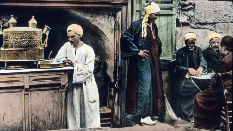 A colorized historical photograph showing a group of men in traditional Middle Eastern attire, including robes and turbans, gathered at an outdoor coffee house. On the left, a man in a white robe stands behind a counter featuring a large, ornate metal coffee urn. To the right, several other men are seated around a small table, engaged in conversation, reflecting the long history of coffee as a social staple.