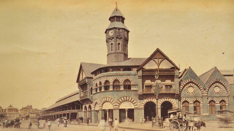 An 1870 sepia-toned photograph of Crawford Market in Mumbai, featuring its prominent stone clock tower and Gothic-style architecture with arched windows. In the foreground, people walk along a dirt road near a horse-drawn carriage and a vintage street lamp.