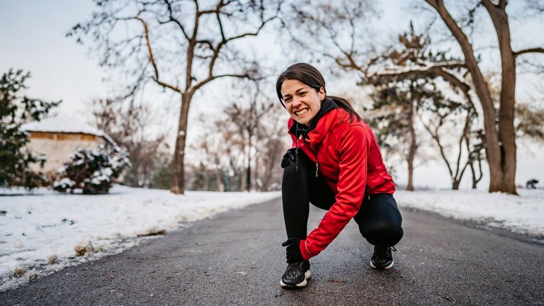 A photograph of a woman in a red jacket outdoors in a snowy park, grimacing in pain while clutching her knee, illustrating how cold weather can affect joint health.