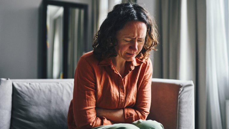 A woman seated indoors, holding her stomach in discomfort and expressing physical distress. The mood captures a realistic depiction of personal pain or health-related issues with natural lighting.